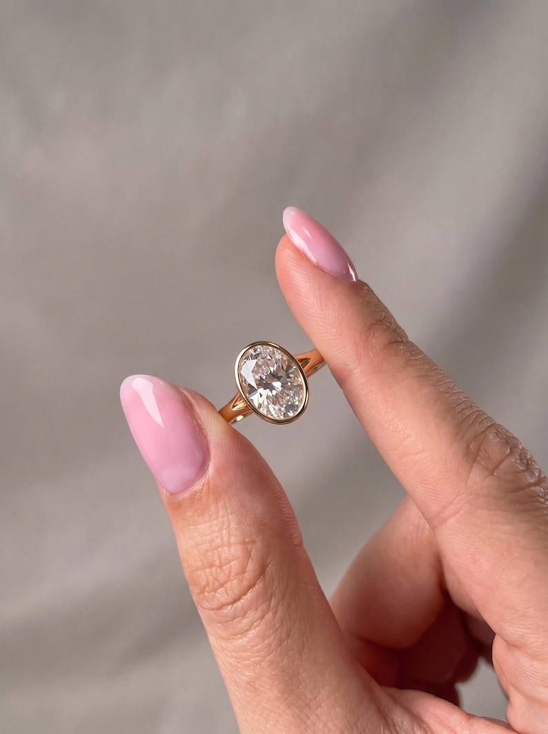 May include: A gold ring featuring a large, clear, oval-cut gemstone set in a smooth, polished bezel setting. The band is thin and circular, crafted from a warm-toned yellow metal that complements the faceted, brilliant-cut center stone.