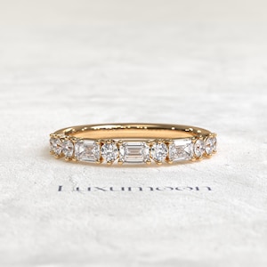 Puede incluir: Un anillo de banda de oro con piedras preciosas claras redondas y de talla esmeralda alternadas. El anillo está sobre una superficie blanca. Una joya delicada.