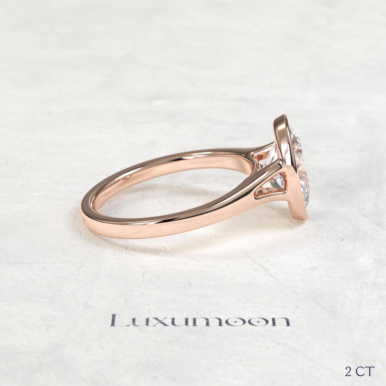 May include: A rose gold ring featuring a clear, pear-shaped gemstone set in a bezel setting. The band is smooth and polished. The text Luxumoon is printed below the ring, with the text 2 CT positioned in the bottom right corner of the image.