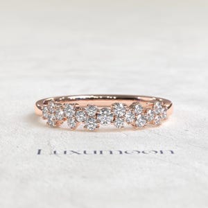 May include: A rose gold ring featuring a cluster of multiple round brilliant-cut clear stones arranged in a scattered, organic pattern across the top of the band. The word Luxumoon is printed in dark blue text below the ring on the surface.