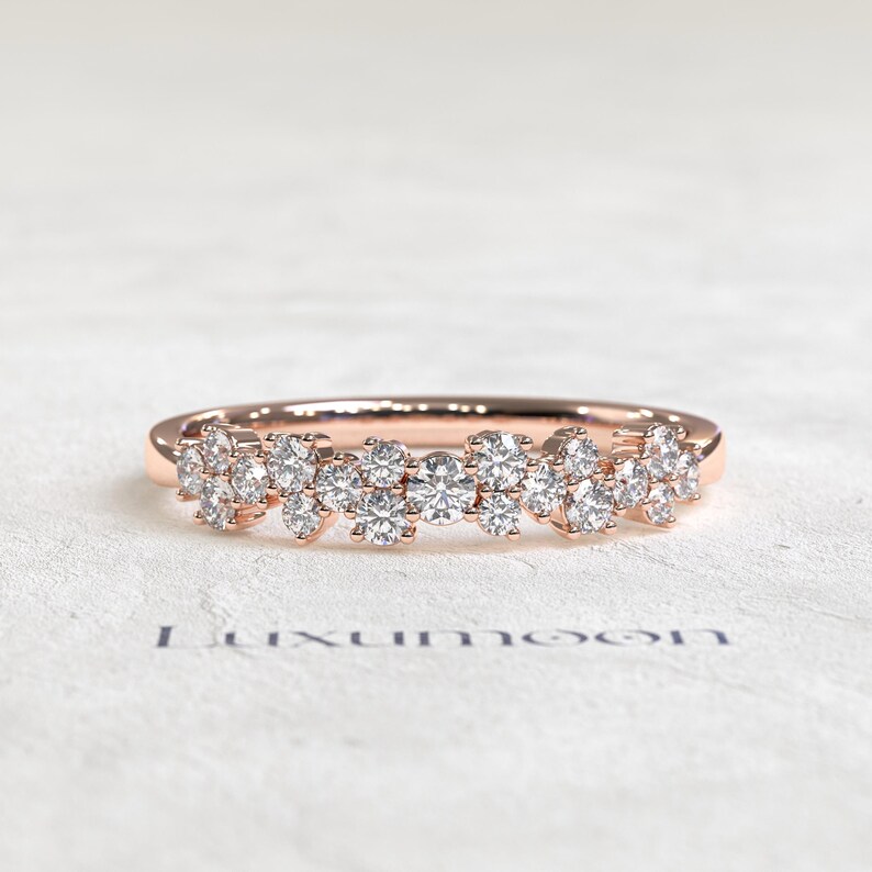 May include: A rose gold ring featuring a cluster of multiple round brilliant-cut clear stones arranged in a scattered, organic pattern across the top of the band. The word Luxumoon is printed in dark blue text below the ring on the surface.
