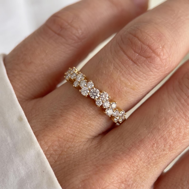 May include: A gold band ring featuring a cluster of small, round, clear, faceted stones arranged in a staggered, multi-layered pattern across the top surface, creating a textured, sparkling appearance on the finger.