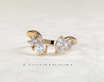 May include: A gold open ring featuring a cluster of clear stones on each end. One side displays a princess-cut stone and a round brilliant-cut stone, while the other side features a pear-shaped stone and a smaller round stone. The text Luxumoon is visible.