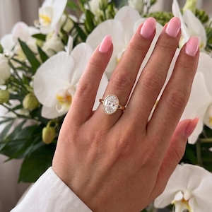 May include: A gold ring featuring a large, oval-cut clear gemstone set in a smooth bezel setting. The band is thin and polished, showcasing a classic design that highlights the clarity and brilliance of the central oval stone on a simple metal band.