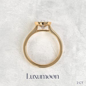 May include: A gold ring featuring a round, clear, faceted gemstone set in a raised, polished gold setting. The band is smooth and circular. The text Luxumoon and 2 CT are printed below the ring, indicating the brand name and a 2 carat weight.