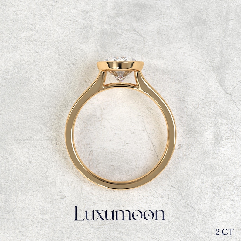May include: A gold ring featuring a round, clear, faceted gemstone set in a raised, polished gold setting. The band is smooth and circular. The text Luxumoon and 2 CT are printed below the ring, indicating the brand name and a 2 carat weight.