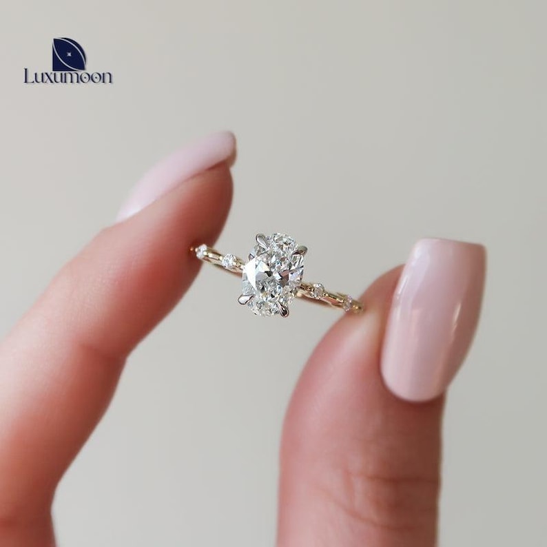 May include: A delicate gold engagement ring featuring a large oval-shaped diamond set in a prong setting. The band is adorned with smaller diamonds that create a delicate vine-like design.