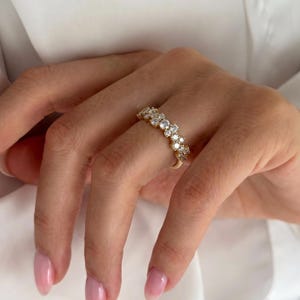May include: A gold band ring featuring a row of multiple round, clear, faceted stones set along the top. The ring has a polished metallic finish and the stones are arranged in a continuous line, creating a sparkling effect across the finger.