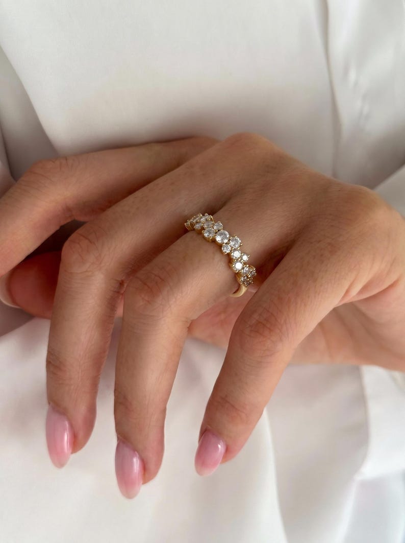 May include: A gold band ring featuring a row of multiple round, clear, faceted stones set along the top. The ring has a polished metallic finish and the stones are arranged in a continuous line, creating a sparkling effect across the finger.