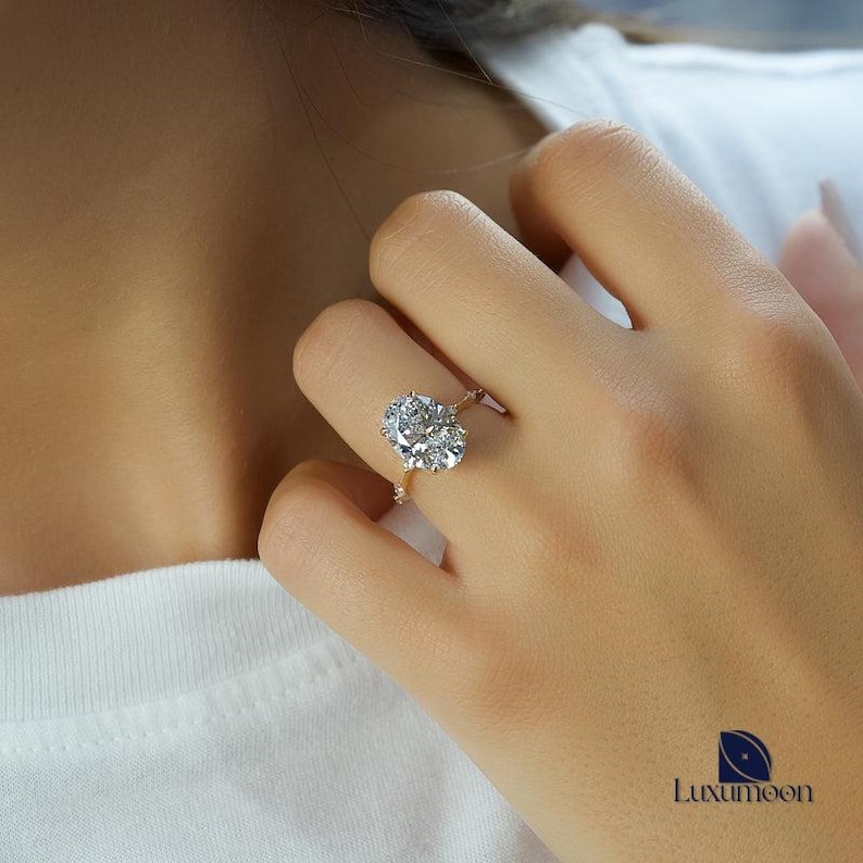 May include: A close-up of a hand wearing a gold ring with a large oval-shaped diamond surrounded by smaller diamonds.