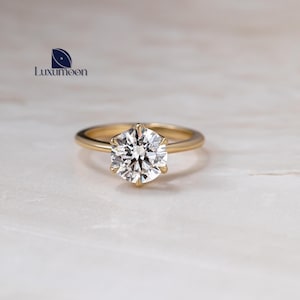 May include: A gold engagement ring featuring a large, round, clear diamond. The diamond is held in place by a six-prong setting. The ring is set against a white and beige marble surface. The brand name "Luxumoon" is visible.