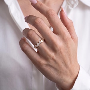 May include: A gold band ring featuring a continuous row of small, round, clear, faceted stones set in a cluster pattern. The ring is worn on a finger, showcasing the metallic gold finish and the sparkling, reflective quality of the stones.