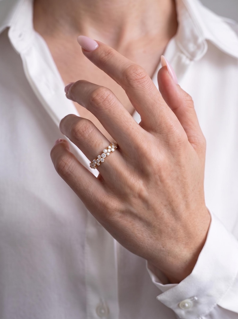 May include: A gold band ring featuring a continuous row of small, round, clear, faceted stones set in a cluster pattern. The ring is worn on a finger, showcasing the metallic gold finish and the sparkling, reflective quality of the stones.