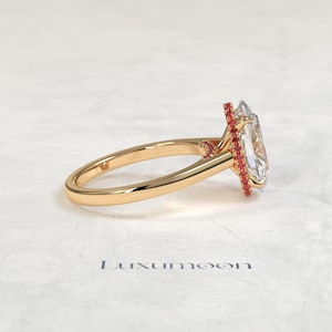 May include: A gold ring with a large, clear, rectangular-cut gemstone. The gemstone is surrounded by a halo of small, red stones. The ring band is smooth and polished. The word "Luxumoon" is printed on the surface.