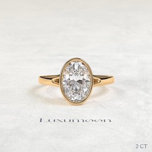 May include: A gold ring featuring a large oval-cut clear gemstone set in a smooth bezel setting. The band has a split shank design that connects to the central stone. The text Luxumoon and 2 CT are visible below the ring on the surface.