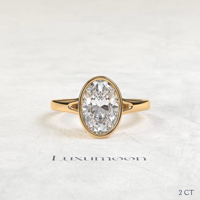 May include: A gold ring featuring a large oval-cut clear gemstone set in a smooth bezel setting. The band has a split shank design that connects to the central stone. The text Luxumoon and 2 CT are visible below the ring on the surface.