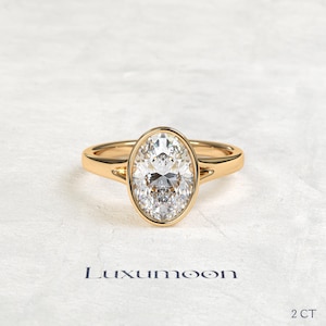 May include: A gold ring featuring a central oval-cut clear gemstone set in a smooth bezel setting. The band is polished gold with a split shank design near the stone. The text Luxumoon and 2 CT are visible at the bottom of the image.