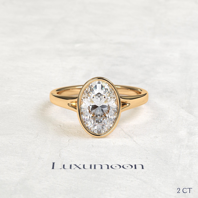 May include: A gold ring featuring a central oval-cut clear gemstone set in a smooth bezel setting. The band is polished gold with a split shank design near the stone. The text Luxumoon and 2 CT are visible at the bottom of the image.