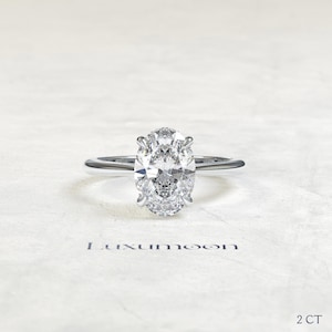 Peut inclure: Une bague de fiançailles solitaire avec un diamant de taille ovale monté sur un fin anneau en argent poli. Le diamant est le point central, brillant. Le texte "Luxumoon" est imprimé en dessous, avec "2 CT" en bas à droite.