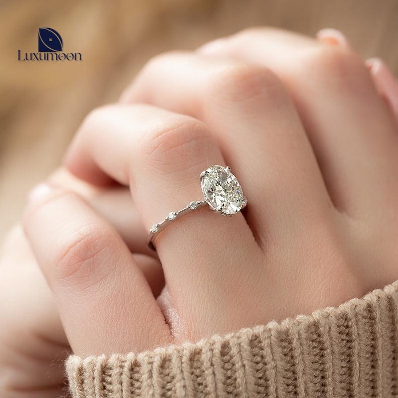 May include: A silver engagement ring featuring an oval-shaped diamond and a delicate band with small diamonds. The ring is worn on a finger.