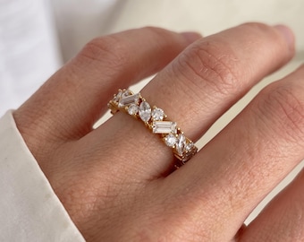 May include: A gold band ring featuring a repeating pattern of alternating clear baguette, marquise, and round cut stones. The stones are set in a continuous row around the circumference of the metal band, creating a textured and reflective surface.