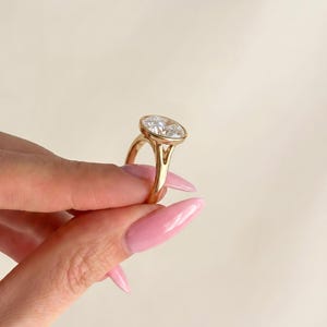 May include: A gold ring featuring a large, clear, oval-shaped faceted gemstone set in a smooth bezel setting. The band has a split shank design that connects to the base of the setting, showcasing a polished metallic finish and a brilliant stone.