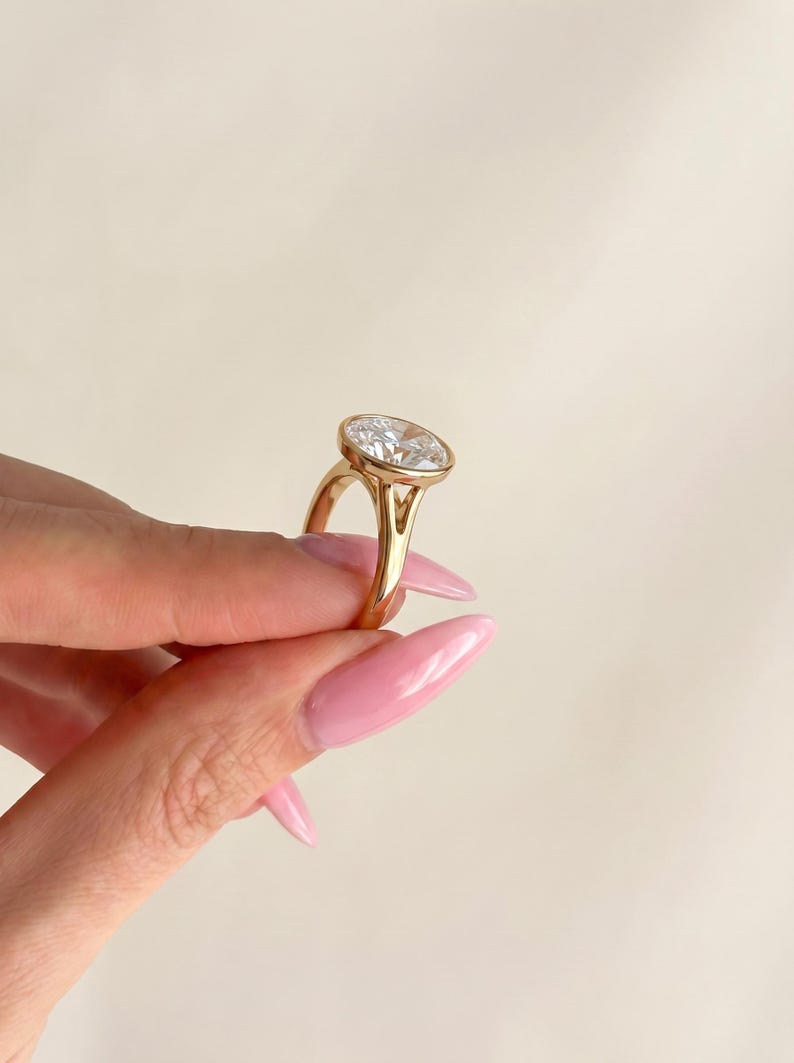 May include: A gold ring featuring a large, clear, oval-shaped faceted gemstone set in a smooth bezel setting. The band has a split shank design that connects to the base of the setting, showcasing a polished metallic finish and a brilliant stone.