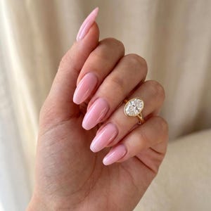 May include: A gold ring featuring a large, oval-cut clear gemstone set in a smooth bezel setting. The ring is worn on the ring finger, which is adorned with glossy, light pink, almond-shaped nails. The band of the ring is thin and polished gold.