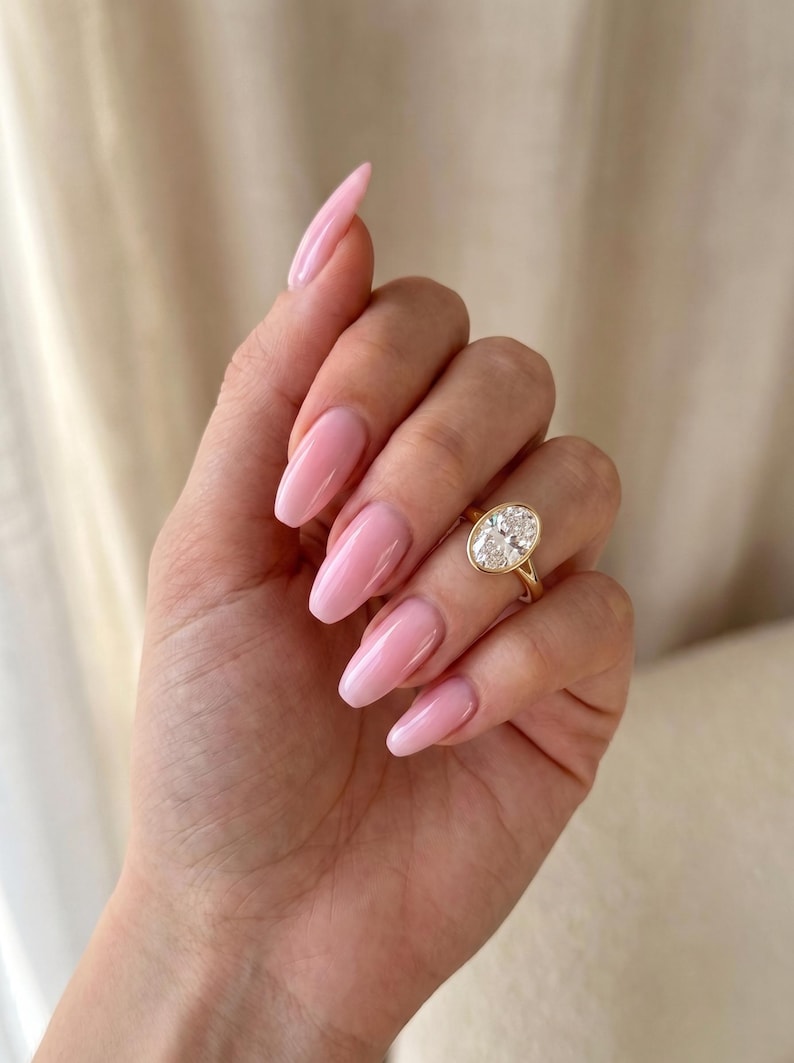 May include: A gold ring featuring a large, oval-cut clear gemstone set in a smooth bezel setting. The ring is worn on the ring finger, which is adorned with glossy, light pink, almond-shaped nails. The band of the ring is thin and polished gold.