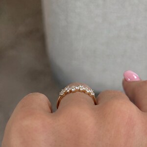 May include: A gold ring featuring a curved band set with a row of small, clear, round-cut stones. The band has a polished metallic finish and the stones are arranged in a cluster formation along the top arc of the ring, creating a sparkling effect.