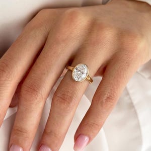 May include: A gold ring featuring a large, clear, oval-cut gemstone set in a smooth bezel setting. The band is thin and polished, showcasing a classic design that highlights the brilliance of the central stone against the warm yellow gold metal.