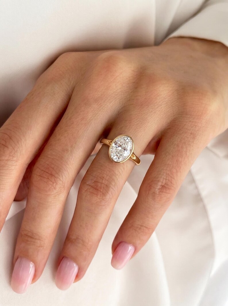 May include: A gold ring featuring a large, clear, oval-cut gemstone set in a smooth bezel setting. The band is thin and polished, showcasing a classic design that highlights the brilliance of the central stone against the warm yellow gold metal.