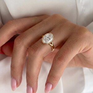May include: A gold ring featuring a large, oval-cut clear gemstone set in a smooth bezel setting. The band is crafted from polished yellow gold and features a split shank design where the band meets the central oval stone setting.