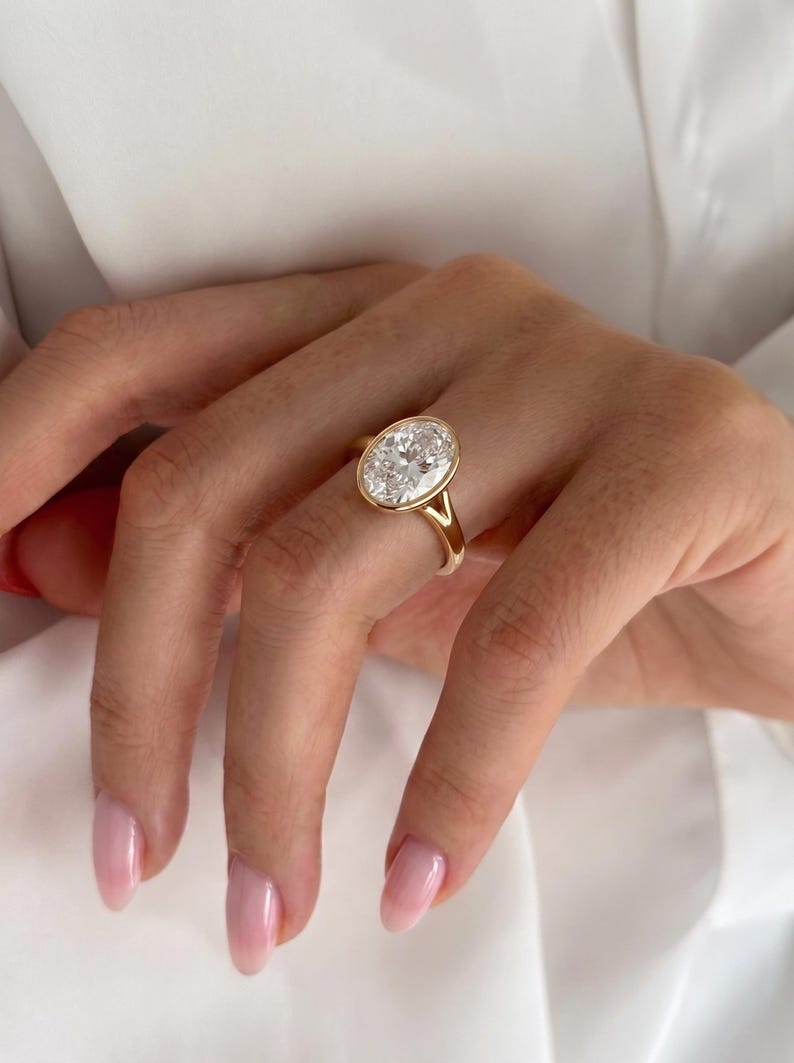 May include: A gold ring featuring a large, oval-cut clear gemstone set in a smooth bezel setting. The band is crafted from polished yellow gold and features a split shank design where the band meets the central oval stone setting.