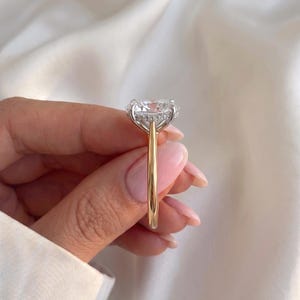 May include: A ring featuring a large, clear, cushion-cut centre stone set in a silver-toned prong setting with a hidden halo of small pavé stones. The band is crafted from polished yellow gold, creating a two-tone metal design with a classic profile.