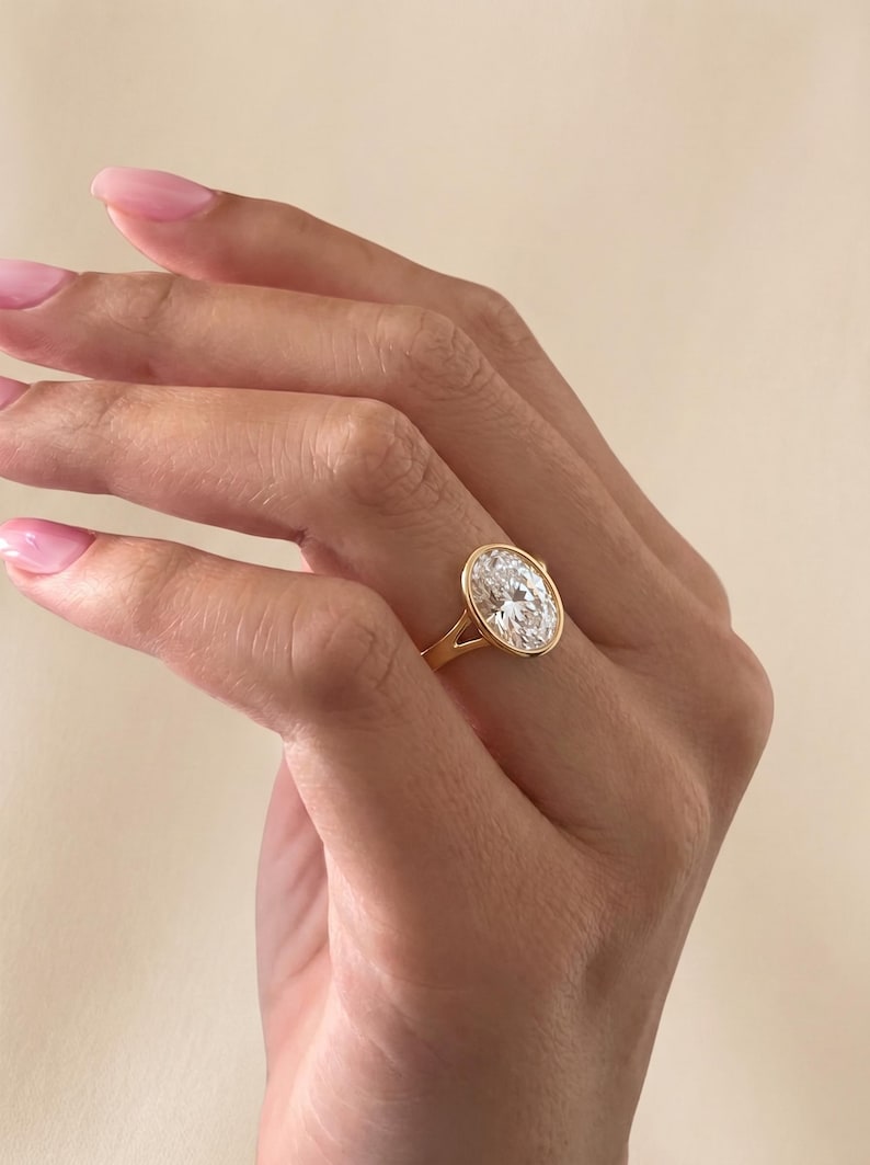 May include: A gold ring featuring a large, oval-cut clear gemstone set in a smooth bezel setting. The band is crafted from polished yellow gold and features a split shank design that connects to the central stone setting, creating a clean look.