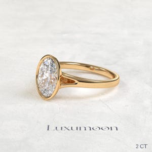 May include: A gold ring featuring a large oval-cut clear gemstone set in a smooth bezel setting. The band has a split shank design where it meets the stone. The text Luxumoon and 2 CT are printed below the ring on the surface.