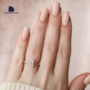 May include: A delicate oval-shaped diamond engagement ring with a halo setting and a band adorned with smaller diamonds. The ring is on a hand with light pink nail polish.