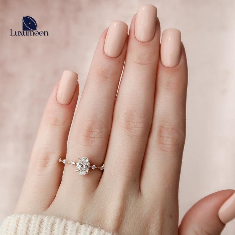 May include: A delicate oval-shaped diamond engagement ring with a halo setting and a band adorned with smaller diamonds. The ring is on a hand with light pink nail polish.