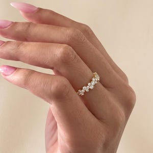 May include: A gold ring featuring a cluster of small, round, clear stones arranged in a continuous, slightly irregular band. The stones are set in a metallic gold frame that follows the contour of the stones, creating a textured, sparkling appearance.