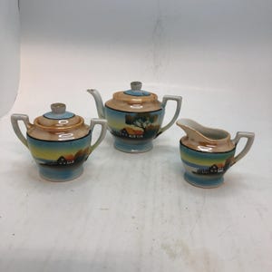 May include: A vintage porcelain tea set featuring a teapot, sugar bowl, and creamer. Each piece is painted with a landscape scene of a house, tree, and sunset in shades of blue, orange, and yellow. The set has a light blue base.