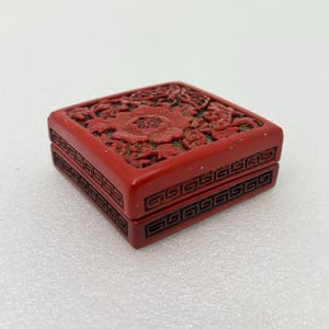 May include: A square red box featuring intricate carved floral patterns on the lid and a geometric Greek key border design along the sides. The box has a deep red finish with darker red accents within the recessed areas of the detailed carvings.