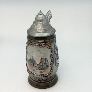 May include: A ceramic beer stein with a metallic lid featuring an embossed dome design. The body is grey with a central rectangular panel depicting a town scene with buildings and the text "STEIN AM RHEIN" framed by a decorative gold-toned border.