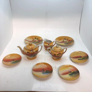 May include: A porcelain tea set featuring a teapot, a lidded sugar bowl, a creamer, and five saucers. Each piece displays a hand-painted landscape with orange, yellow, and brown hues, depicting a windmill near water under a sunset sky.