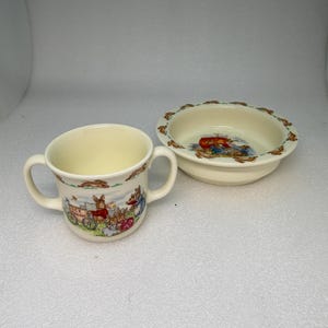 May include: A cream-colored ceramic children's cup with two handles and a matching bowl. Both pieces feature a colorful illustration of rabbits and a small ice cream cart. The bowl has a scalloped edge with rabbit designs.