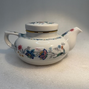 May include: A white porcelain teapot featuring a delicate floral pattern with blue, pink, and orange blossoms. The teapot has a matching lid with a gold-tone rim, a curved handle, and a short spout, all decorated with fine botanical motifs.