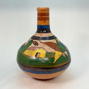 May include: A hand-painted ceramic vase with a rounded body and narrow neck. The surface features a colorful landscape scene with a yellow house, green foliage, and brown accents. Horizontal stripes in orange, blue, and black decorate the neck. Mexico is written.