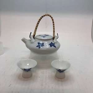 May include: A white ceramic teapot with a blue maple leaf pattern and a woven bamboo handle, accompanied by two matching small ceramic cups. The teapot lid features blue leaf motifs and black Japanese characters, while the cups have a flared rim.