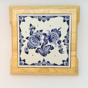 May include: A square ceramic tile featuring a blue and white floral pattern, framed in light-colored natural wood. The tile displays intricate blue botanical illustrations on a white background, with a decorative border of blue floral motifs.