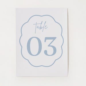 May include: A rectangular white table number card featuring a light blue scalloped border. Centered within the border is the word Table in a cursive script font, positioned above the large, bold, light blue number 03.
